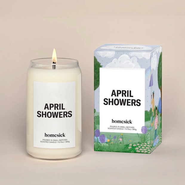 A lit April Showers Homesick candle displayed next to its boxed packaging on a dark cream background.