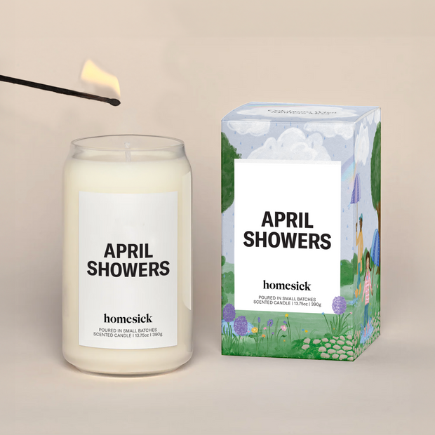 A lit April Showers Homesick candle displayed next to its boxed packaging on a dark cream background.