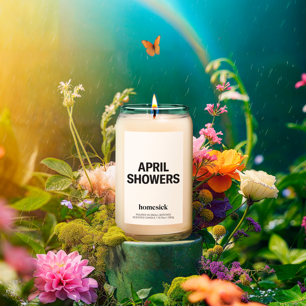 An April Showers Homesick candle set on a pedestal surrounded by flowers and native plants and grasses, set on the foreground of sprinkling rain and a rainbow