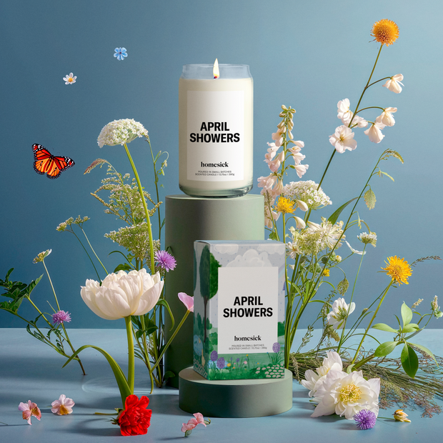 An April Showers candle on a high pedestal set behind its boxed packaging on a shallower pedestal, surrounded by flowers native plants and grasses, a small vibrant monarch butterfly, on a powder blue gradient background.