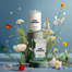 An April Showers candle on a high pedestal set behind its boxed packaging on a shallower pedestal, surrounded by flowers native plants and grasses, a small vibrant monarch butterfly, on a powder blue gradient background.