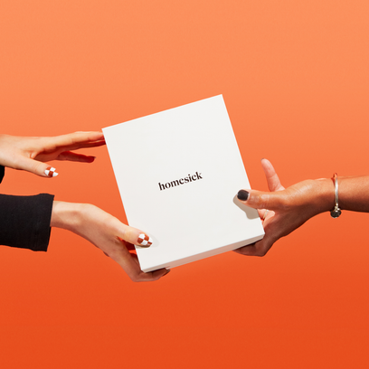 A hand passing to another hand a box of Homesick candles. The hands are in front of a bright orange background.