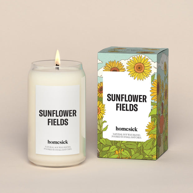 A lit Sunflower Fields Homesick candle displayed next to its boxed packaging on a dark cream background.