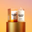 A lit Golden Hour Homesick candle displayed next to its boxed packaging on a golden pedestal in front of a vibrant background.