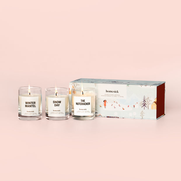 All three lit Winter Wonderland Mini Candles next to their boxed packaging on a dark cream background.