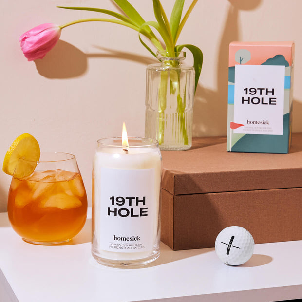 A lifestyle image of the 19th Hole Homesick candle with various golf props surrounding it.