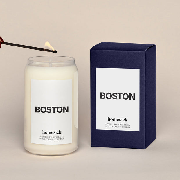A lit Boston Homesick candle displayed next to its boxed packaging on a dark cream background.