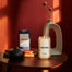 A lifestyle image of the Boston Homesick Candle on a side table with various home decor also on the table. There is a red background and lighting is moody.