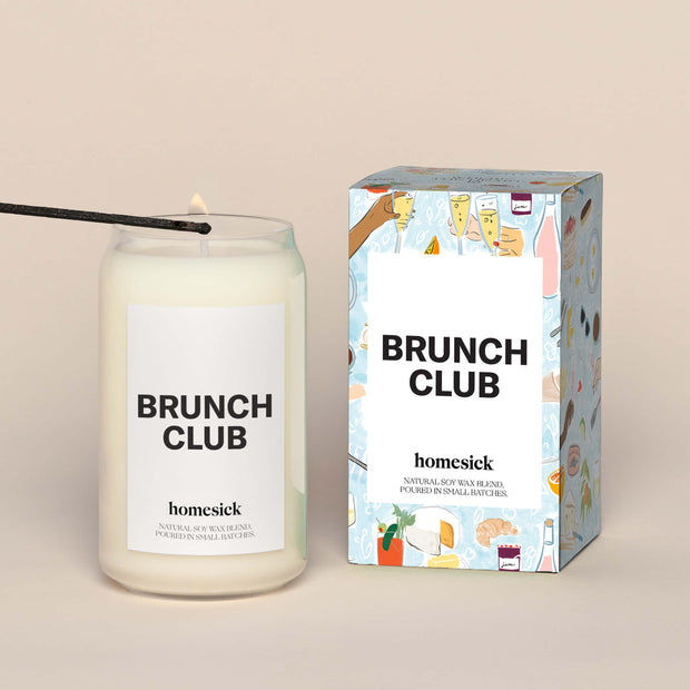 A lit Brunch Club Homesick candle displayed next to its boxed packaging on a dark cream background.