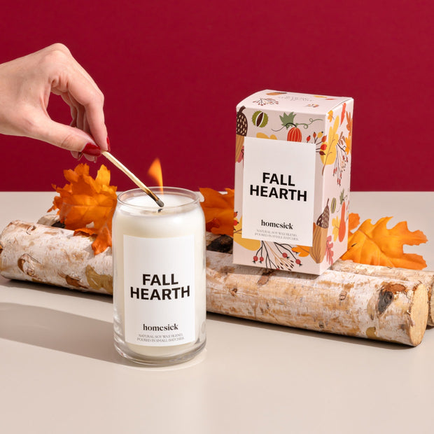 Someone lighting the Fall Hearth Candle that is on a light gray surface. Behind it are two aspen logs where the candle packaging sits on top of.