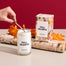 Someone lighting the Fall Hearth Candle that is on a light gray surface. Behind it are two aspen logs where the candle packaging sits on top of.