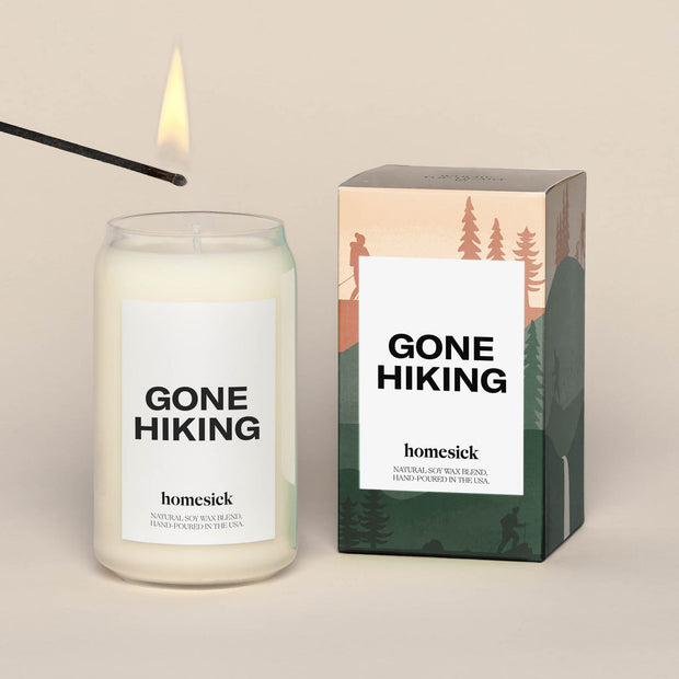Gone Hiking candle placed next to the packing box. The box in a printed scene of mountain and pine tress.