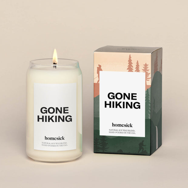 Gone Hiking candle placed next to the packing box. The box in a printed scene of mountain and pine tress.