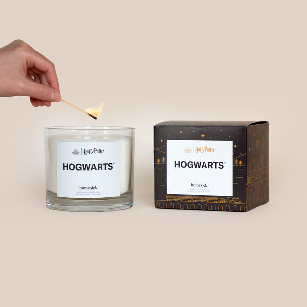 A lit 3-Wick Hogwarts Homesick candle displayed next to its boxed packaging on a dark cream background.
