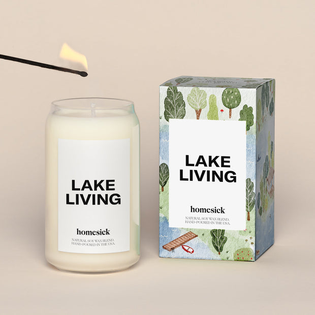 A lit Lake Living Homesick candle displayed next to its boxed packaging on a dark cream background.