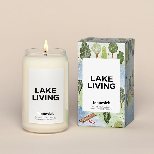 A lit Lake Living Homesick candle displayed next to its boxed packaging on a dark cream background.