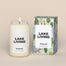 A lit Lake Living Homesick candle displayed next to its boxed packaging on a dark cream background.