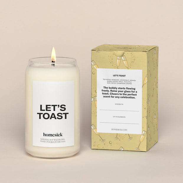 Single Lets Toast Candles next to a the gold box with animated Champagne glasses over all print. There is a white label that reads "Lets Toast"