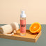 A stylized shot of the Powder Room Linen Spray inside a bamboo tray next to a loofah and cut open orange.