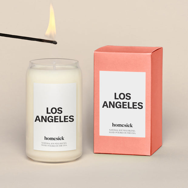A lit Los Angeles Homesick candle displayed next to its boxed packaging on a dark cream background.