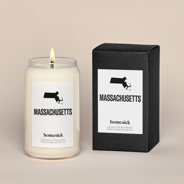A lit Massachusetts Homesick candle displayed next to its boxed packaging on a dark cream background.