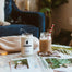 A lifestyle shot inside someone's living room where there is a recently blown out Mississippi Candle next to an iced coffee in a mason jar. Those items are on a white marble table with other personal items like a blanket and magazine.