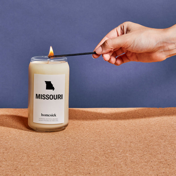 A close up of a Missouri candle displayed on a cork board surface with a navy background. There is a hand entering from the left side that is using a match to light the candle.