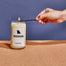 A close up of a Missouri candle displayed on a cork board surface with a navy background. There is a hand entering from the left side that is using a match to light the candle.