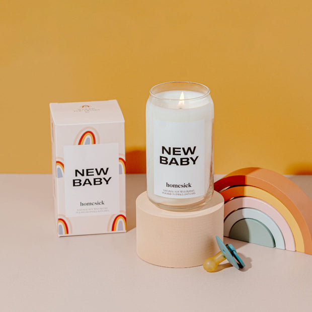 A stylized studio shot of the New Baby candle displayed on a cream rounded pedestal. To the left is the box packaging for the candle with a wooden rainbow toy on the right.