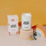 A stylized studio shot of the New Baby candle displayed on a cream rounded pedestal. To the left is the box packaging for the candle with a wooden rainbow toy on the right.