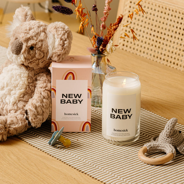 A stylized shot of the New Baby candle and its packaging on a wavy display mat. Next to those are a handful of baby items like a soft koala animal, binky and teether.