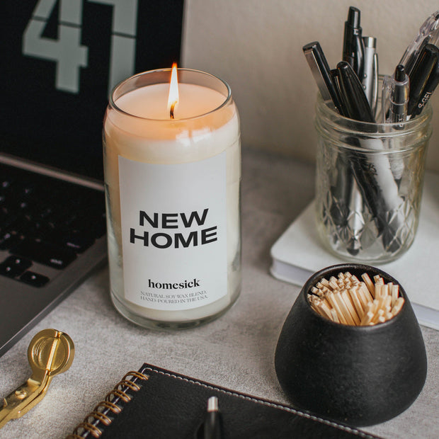 A lit New Home candle sitting next to a laptop, jar of pens and a jar of matches.