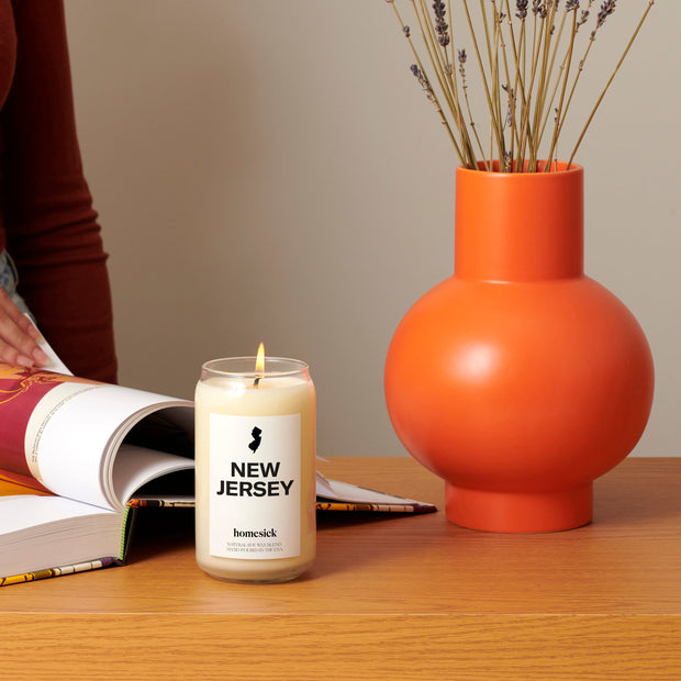 A lifestyle image showing the New Jersey candle in front of someone reading a book. There is a bright orange vase with dried flowers on the right side of the image.