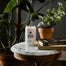 The New York Candle displayed on a rounded marble table that has various books, matches and potted plants also on the table.