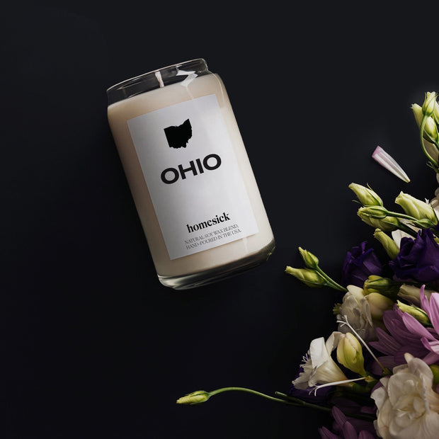 An overhead shot of the Ohio Homesick candle on a jet black background. In the bottom right corner are green and purple fresh flowers.
