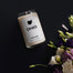 An overhead shot of the Ohio Homesick candle on a jet black background. In the bottom right corner are green and purple fresh flowers.