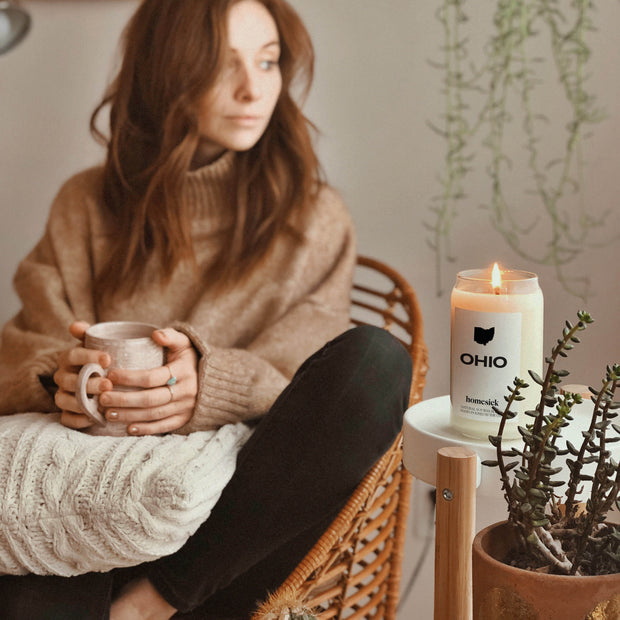 A lifestyle image of the Ohio candle on a white round table. In the blurred background is a woman sitting in a chair holding a cup of tea.