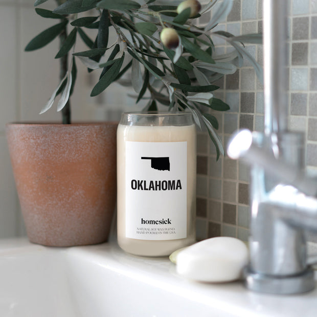 A close up shot of the Oklahoma candle on the edge of a white bathroom sink. There is a cermaic planter with an olive plat next to it.