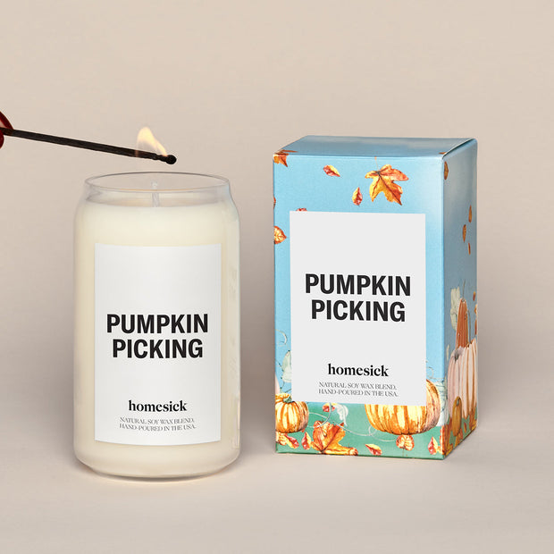 A lit Pumpkin Picking Homesick candle displayed next to its boxed packaging on a dark cream background.