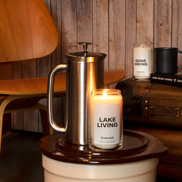 A stylized set of the Lake Living Candle next to a stainless steel french press. in the background are various items once would find in a rustic luxury lake house,