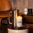 A stylized set of the Lake Living Candle next to a stainless steel french press. in the background are various items once would find in a rustic luxury lake house,