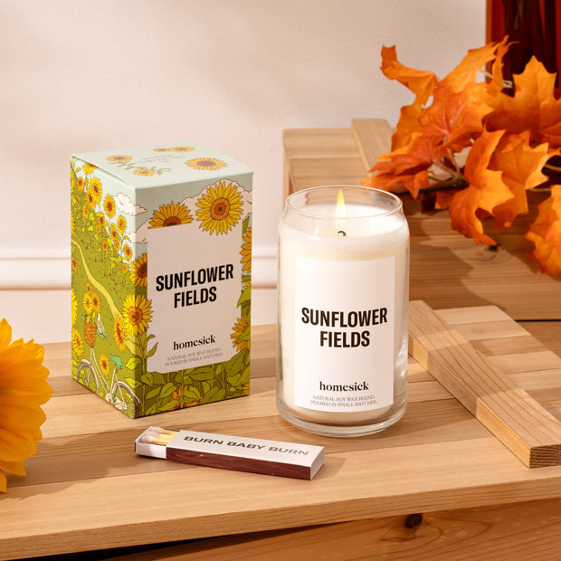 A stylized shot of the Sunflower Fields candle, its packaging, a box of matches on top of a wooden platter. In the back is a light cream wall.