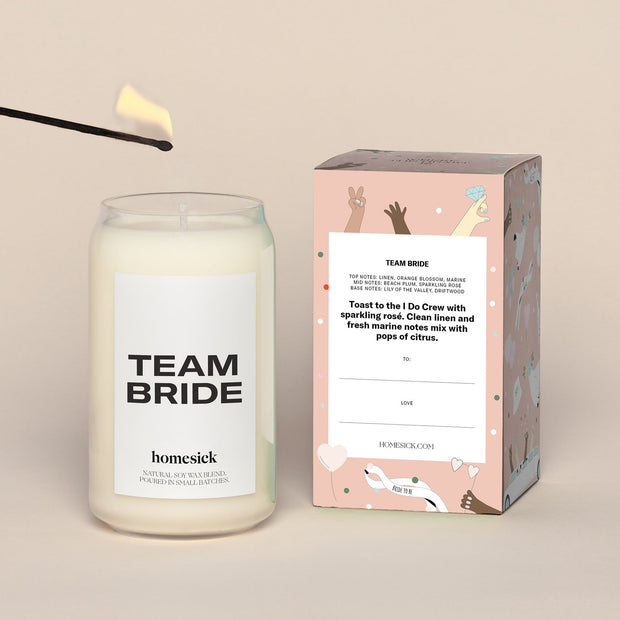 A lit Team Bride Homesick candle displayed next to its boxed packaging on a dark cream background.