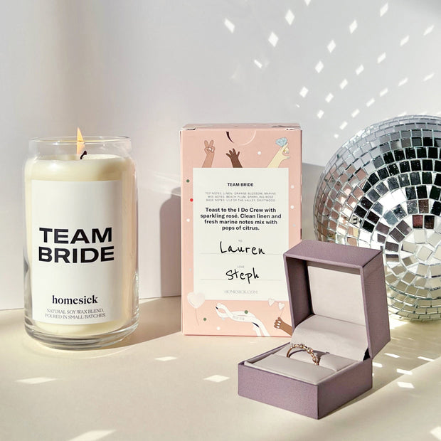 The Team Bride candle on the left, with the back of the packaging in the middle, and a disco ball on the far right. If front of the disco ball is a pale purple ring box.
