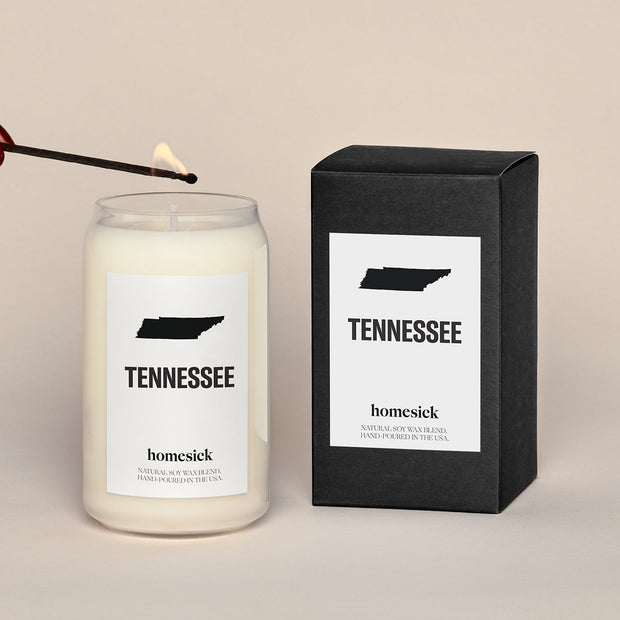 A lit Tennessee Homesick candle displayed next to its boxed packaging on a dark cream background.