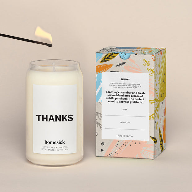 A lit Thanks Homesick candle displayed next to its boxed packaging on a dark cream background.