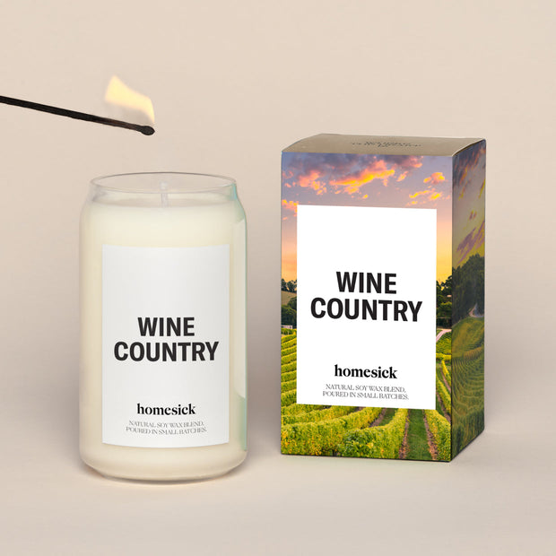 A lit Wine Country Homesick candle displayed next to its boxed packaging on a dark cream background.