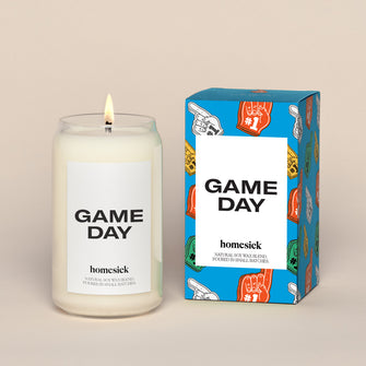 A lit Game day Homesick candle displayed next to its boxed packaging on a dark cream background.