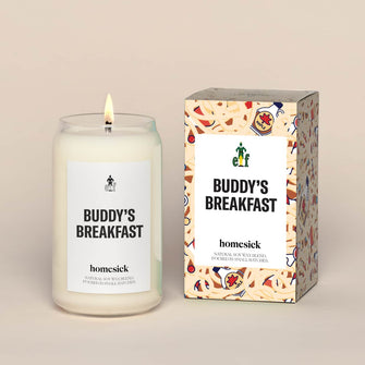 A lit Buddy's Breakfast Homesick candle displayed next to its boxed packaging on a dark cream background.