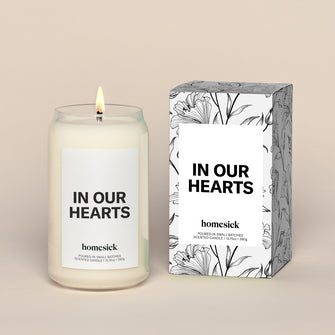 A lit In Our Hearts Homesick candle displayed next to its boxed packaging on a dark cream background.